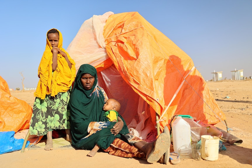 A woman holding a young child sits in front of an orange tent.