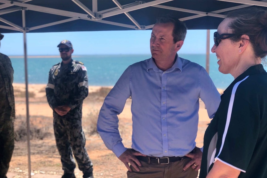 Mark McGowan wearing a blue shirt and brown pants stands with his hands on hips under a blue shade structure.