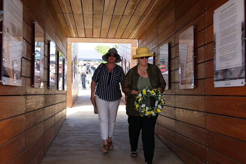 Two women walk through a hall way carrying a flower wreath.