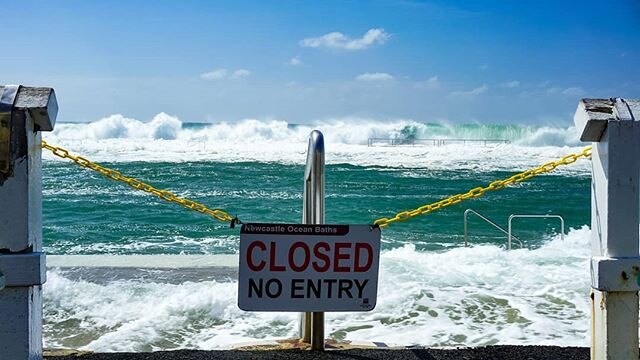 A sign indicating that the Newcastle Ocean Baths are closed, with large waves in the background.