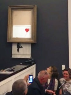 People use mobile phones to film  half-shredded painting of a girl with a red heart-shaped balloon in a big frame on a dark wall