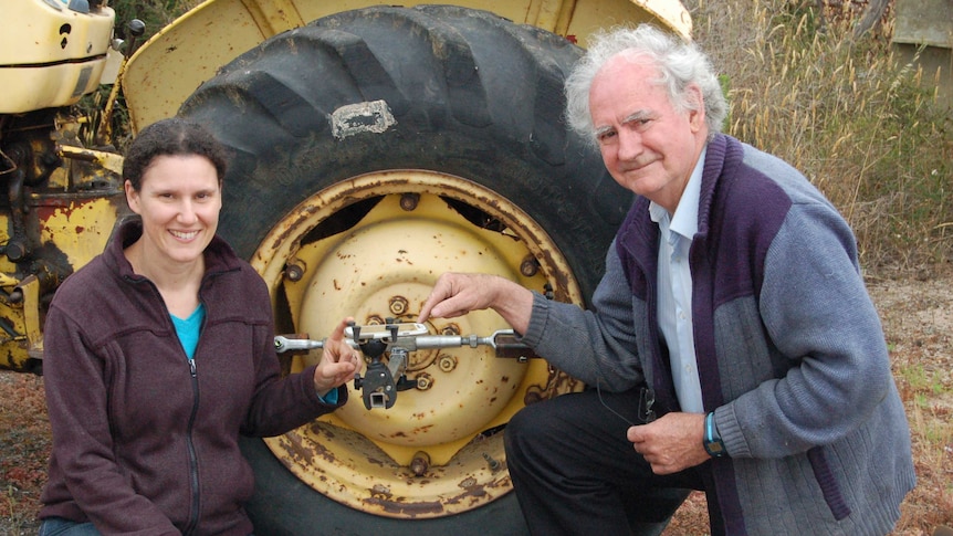 A man and woman squatting beside a tractor wheel with a smartphone clamped to the centre of the wheel.