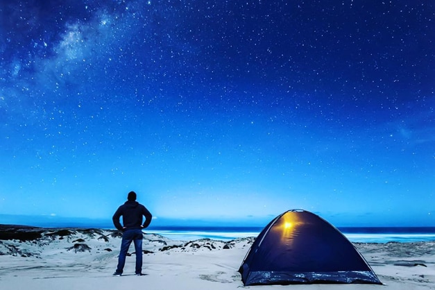 A hiker with a tent pitched next to him looks out at the view of a snowy twilight landscape.