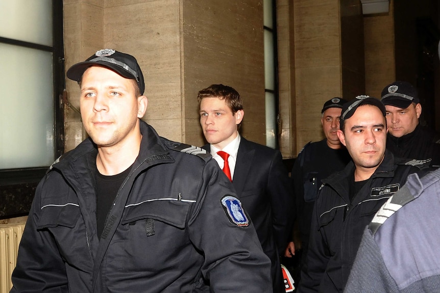 A man wearing a suit and red tie is surrounded by police officers