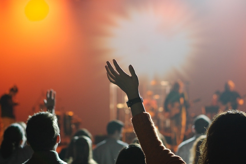 A church congregation, featuring a woman with her hand in the air. A band is playing at the front. Photo taken from the back. 