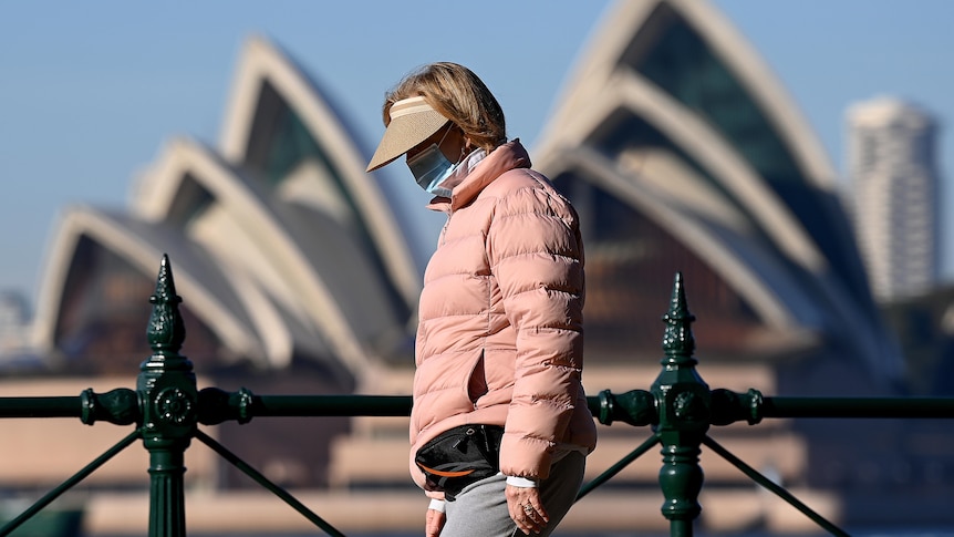 A woman in a pink jacket and peak hat wears a mask with her face in shadow in front of the Sydney Opera House