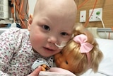 A young girl holds a doll while laying on a hospital bed.