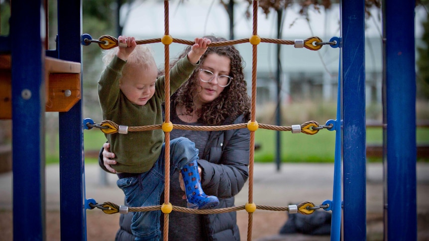 A young white woman carefully lifting a toddler up at a playground