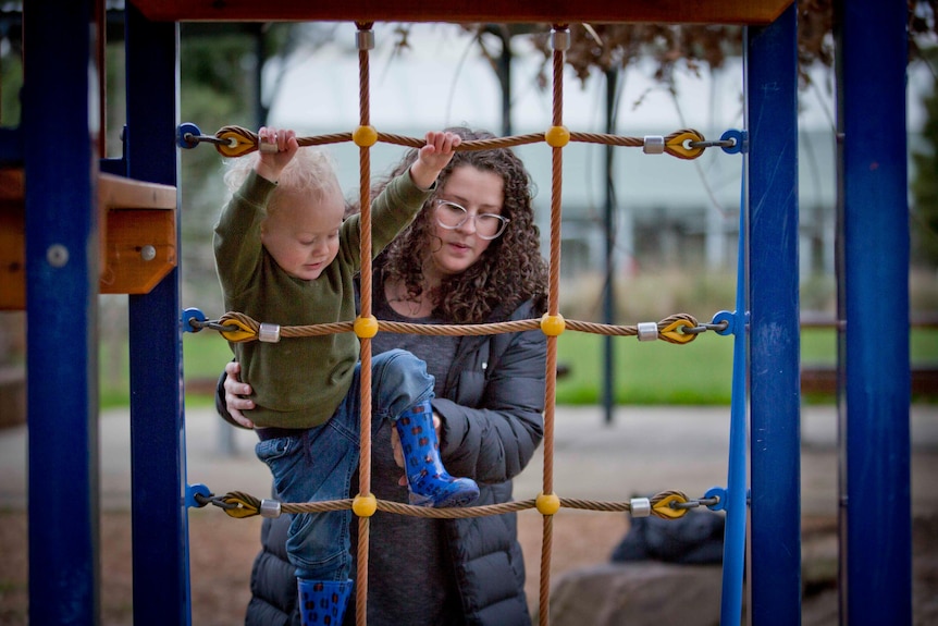 A young white woman carefully lifting a toddler up at a playground