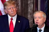 Lindsey Graham speaks at a podium while Donald Trump watches from behind.