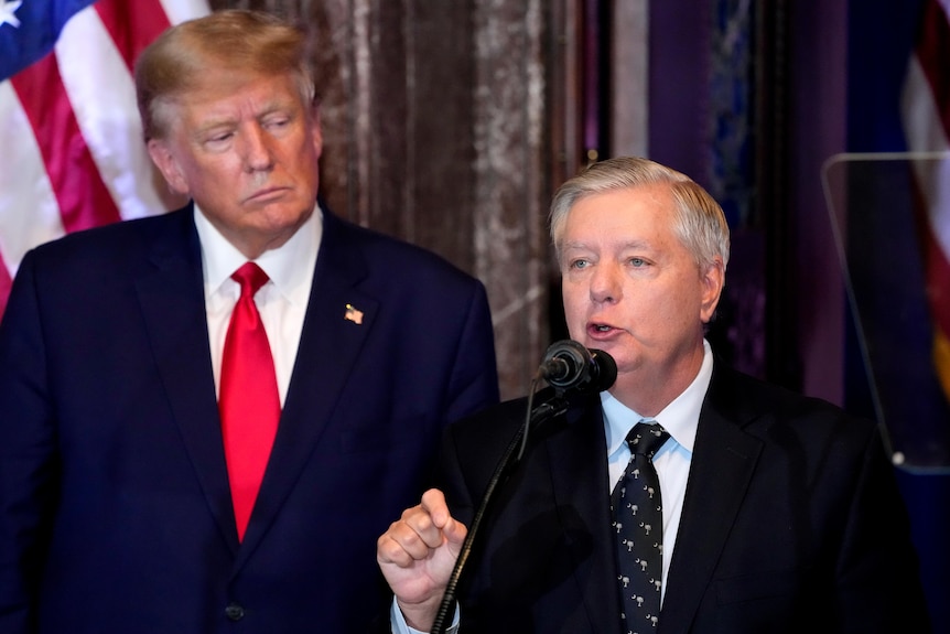 Lindsey Graham speaks at a podium while Donald Trump watches from behind.