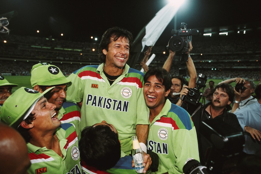 Imran Khan smiles as he is held up by teamates on a cricket pitch.