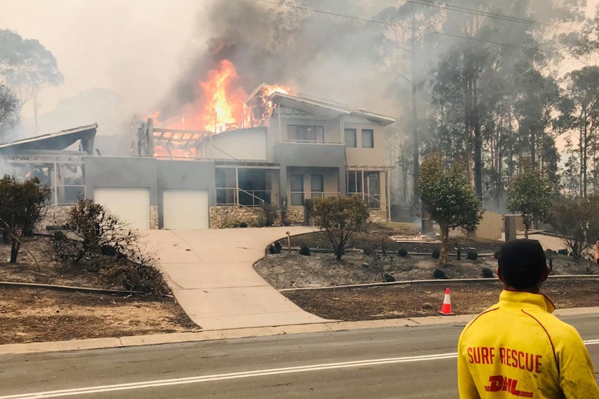 Man in surf lifesaver shirt standing outside a burning house.