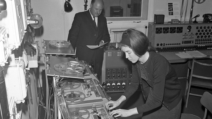 a black and white image of Delia, a young woman in 1960s dress, working at an analogue tape deck, whilst a suited man watches