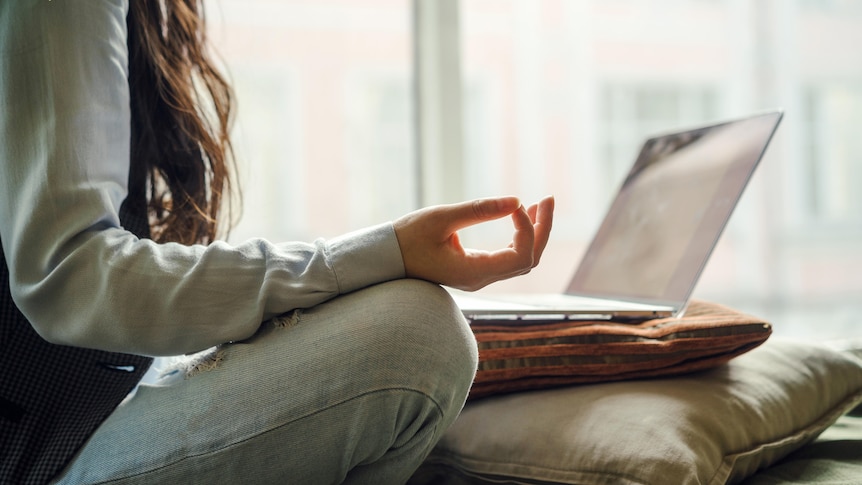 A person with legs crossed sits meditating in front of a laptop on a cushion