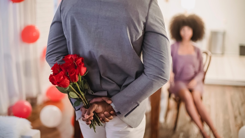 A man holds a bunch of red roses behind his back, wiating to give them to a woman sitting on a chair