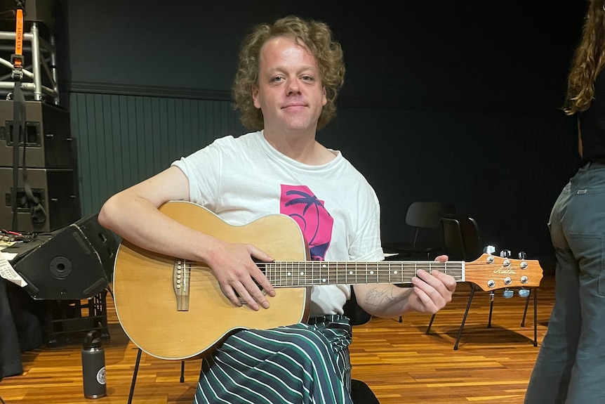 A person with curly hair holds a guitar and looks at the camera. 