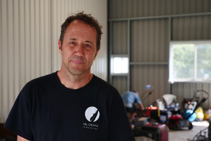 A man with various tools in the background smiles at the camera.