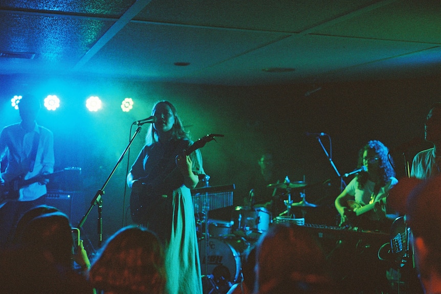 Singer Julia Jacklin stands with guitar on blue light stage with a drummer and guitarist and motel walls behind them