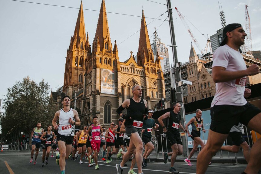 A low view of runners in race along city street with an old church, buildings and trees behind.