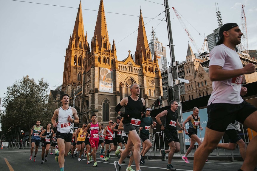 A low view of runners in race along city street with an old church, buildings and trees behind.