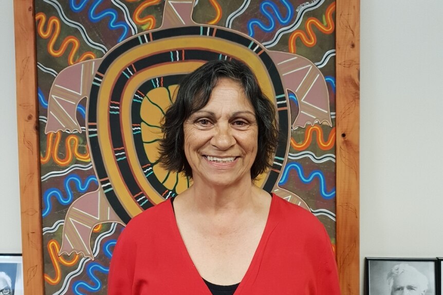 A woman in a red short stands in front of Indigenous artwork 