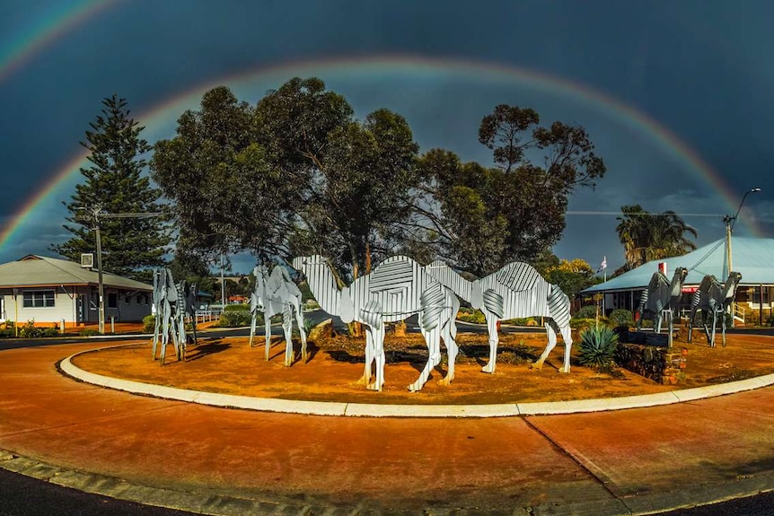 Metal camel sculptures on a roundabout with a rainbow over the top.