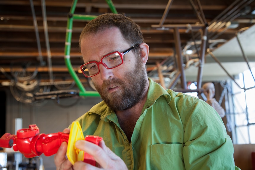 A man in a green collared shirt and red glasses looks down at an object in a busy design lab.