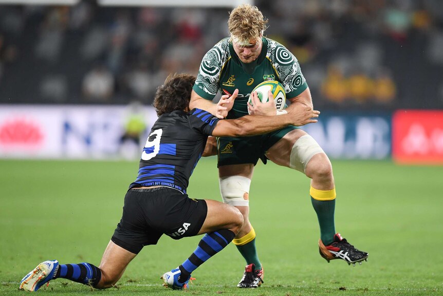 A Wallabies player holds the ball while being tackled by a Pumas opponent.