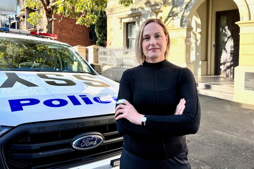 A woman wearing a black top stands next to a police car with her arms crossed.