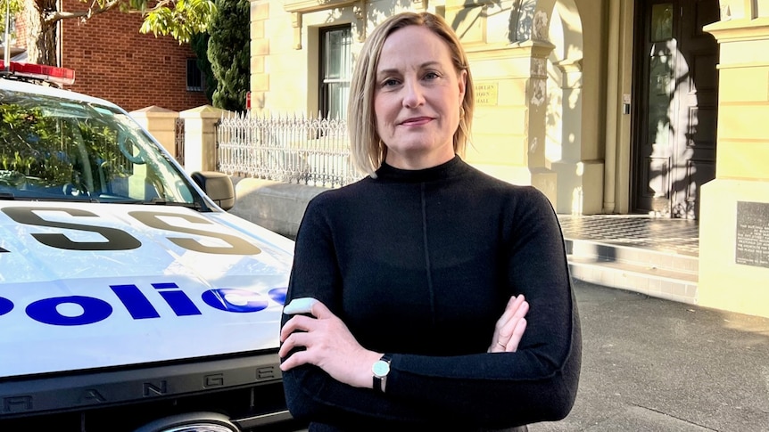 A woman wearing a black top stands next to a police car with her arms crossed.
