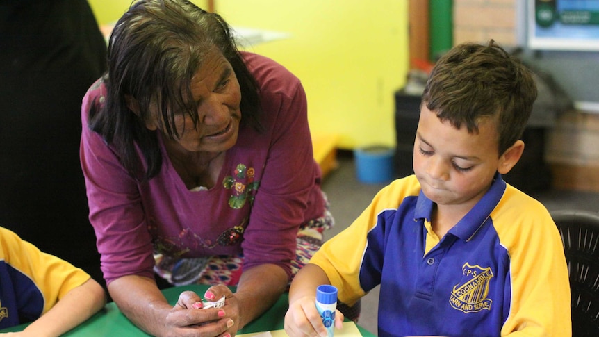 A woman leans over a child's desk to help with activities