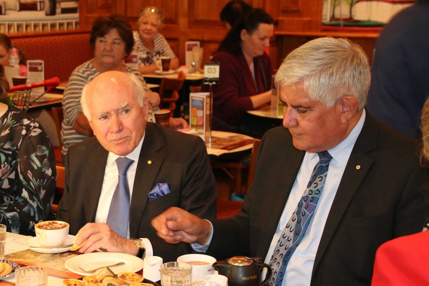 John Howard and Ken Wyatt wearing suits sit at a long table having tea and coffee.