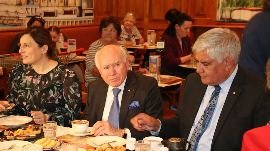 John Howard and Ken Wyatt wearing suits sit at a long table having tea and coffee.
