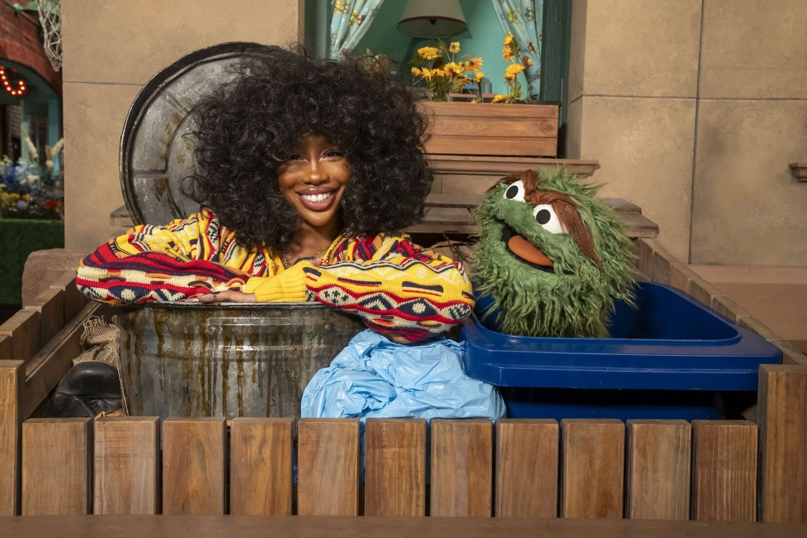 A woman with big curly hair and a green monster sit in trash cans side by side.
