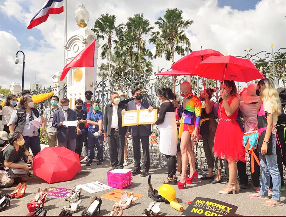 A man holds up a book as people gather around him holding umbrellas and wearing colourful clothes and high heels