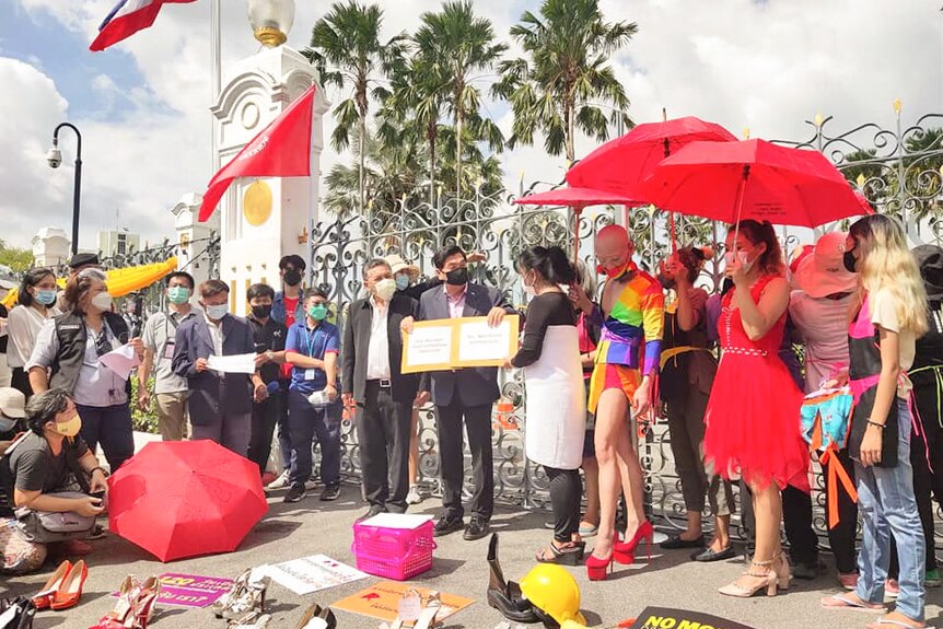 A man holds up a book as people gather around him holding umbrellas and wearing colourful clothes and high heels