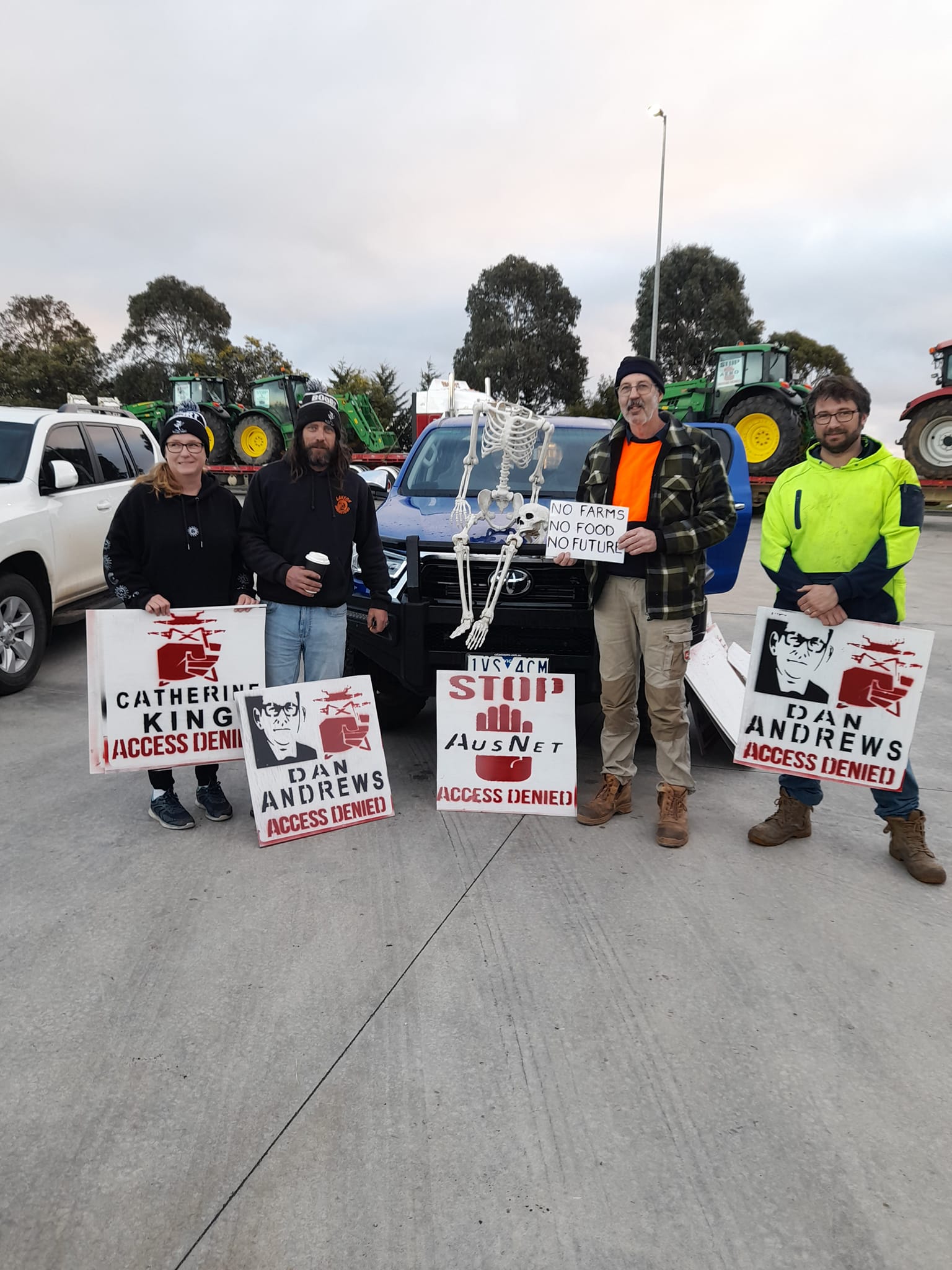 A group of people dressed for warmth stand in front of a truck loaded with tractors. They are holding protest signs.