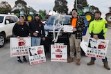 A group of people dressed for warmth stand in front of a truck loaded with tractors. They are holding protest signs.
