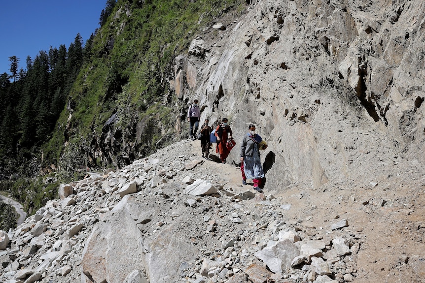A group of doctors walk along a narrow, rocky terrain with a green mountain behind them.