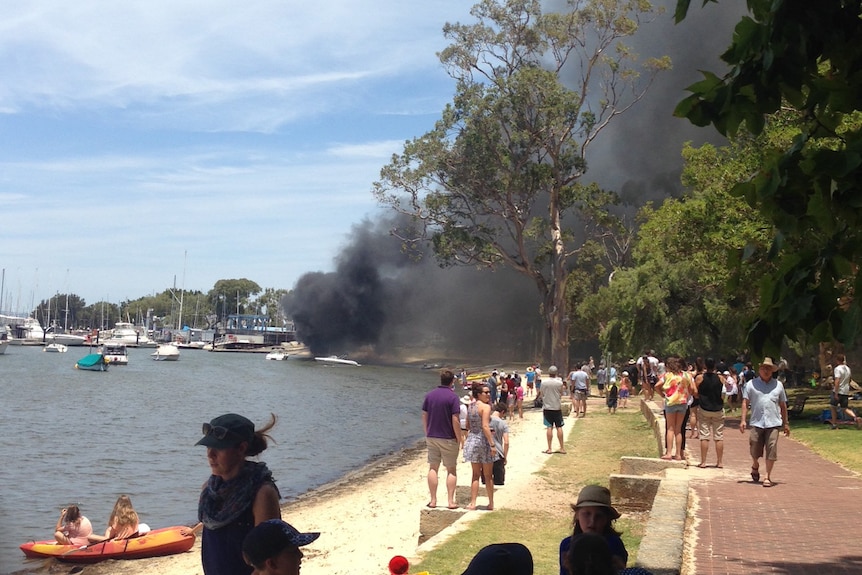 Smoke billows from a boat on fire near the shore of Swan River at Matilda Bay as people watch on.