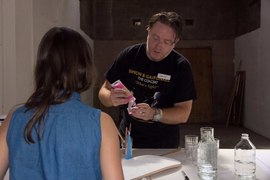 A man squeezes pink paint out of tube with a girl's back in the foreground