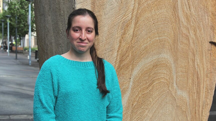 A portrait of Carolina in a green jumper standing against a sandstone wall.