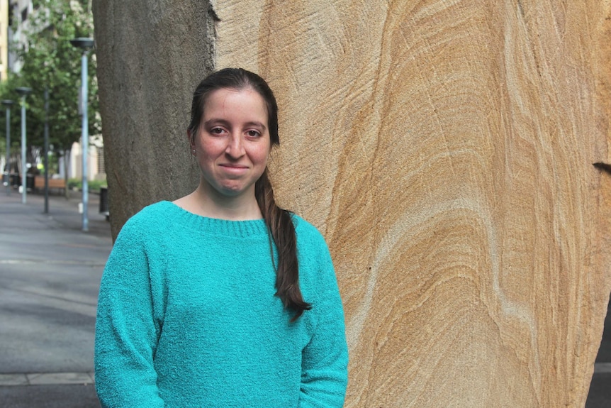 A portrait of Carolina in a green jumper standing against a sandstone wall.