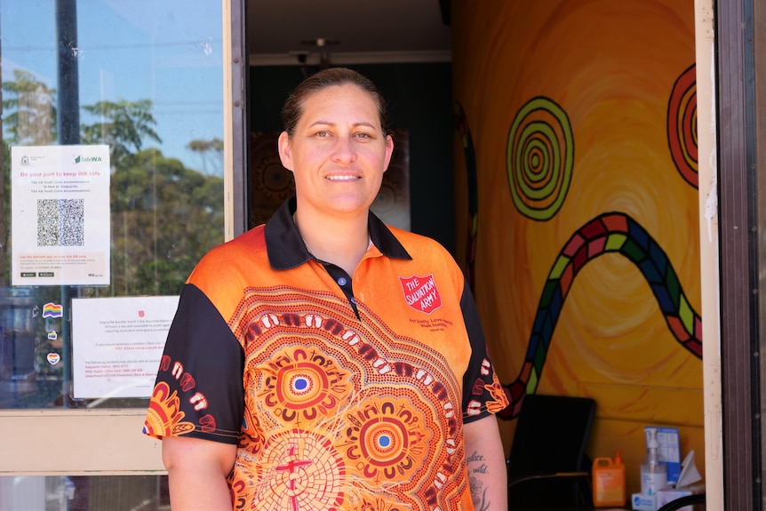 A dark-haired woman in an orange and black t-shirt with Aboriginal motifs stands in front of glass door and an orange wall