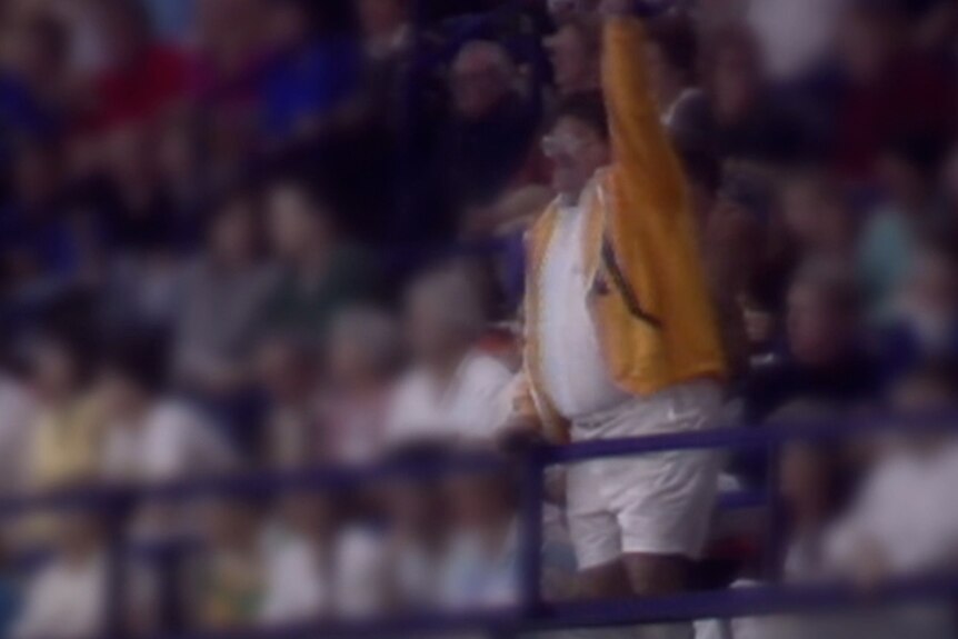 An overweight man in a yellow jacket stands in a crowd cheering.
