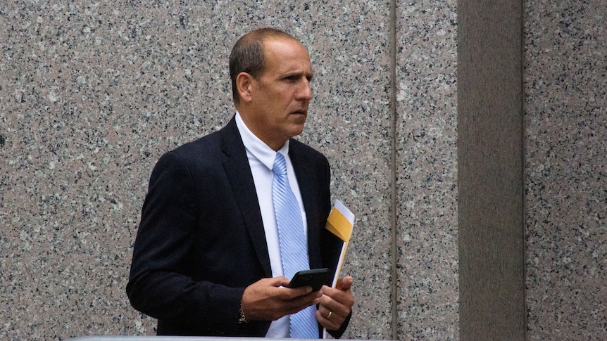 A balding middle aged man stands in front of a wall with a notebook and envelope in hand.