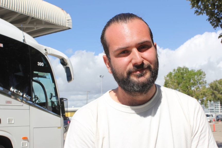 A man in his 20s with a beard, outside in front of a bus.