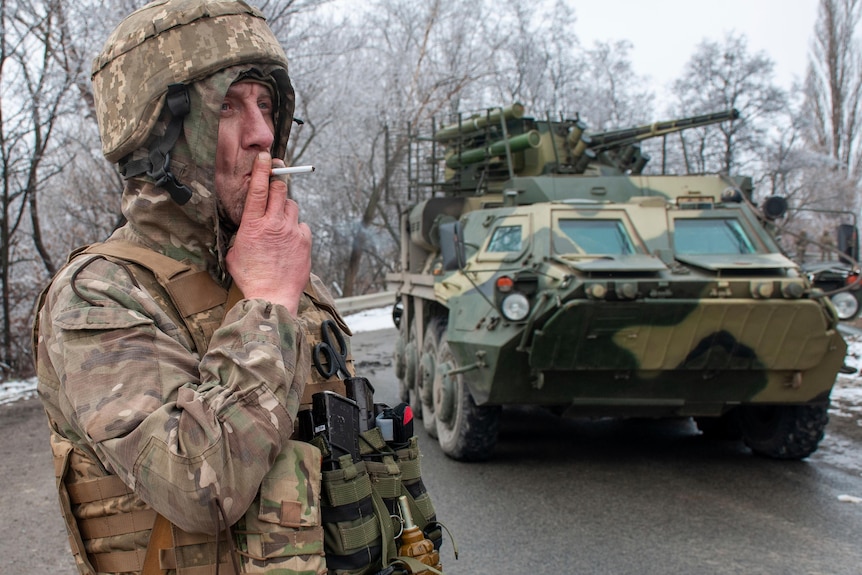 A Ukrainian soldier in military uniform smokes a cigarette, next to a military vehicle.