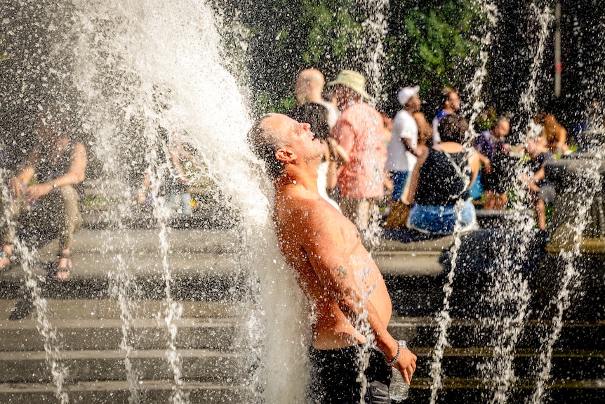 A shirtless man stands in a fountain with his eyes closed 